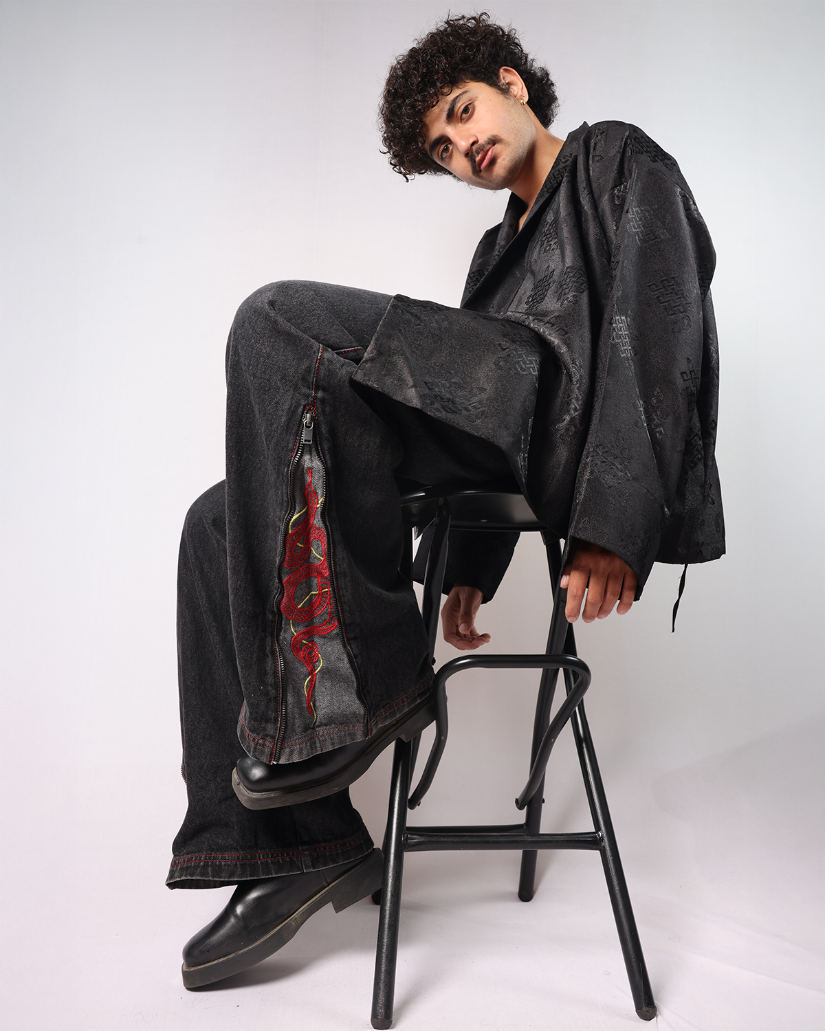 Man sitting on a stool wearing Black Year of Snake jeans and a black jacket, showcasing a stylish look.