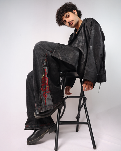 Man sitting on a stool wearing Black Year of Snake jeans and a black jacket, showcasing a stylish look.